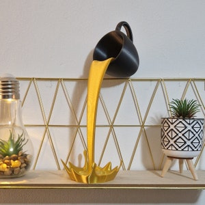 Puede incluir: Un líquido de color dorado que se vierte de una taza de cerámica negra, formando una salpicadura decorativa. La taza está inclinada hacia un lado y el líquido está congelado en el aire. La escultura está sobre un estante con un fondo de malla de alambre dorado.