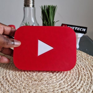 Puede incluir: Un logotipo de YouTube de plástico rojo con un botón de reproducción blanco. El logotipo está sobre una superficie de paja tejida.