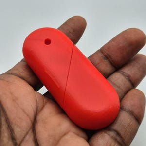 Puede incluir: Un objeto rojo brillante, de forma oblonga, con un peque&ntilde;o agujero en un extremo y una l&iacute;nea diagonal en la superficie. El objeto se sostiene en una mano de piel oscura, sobre un fondo blanco.