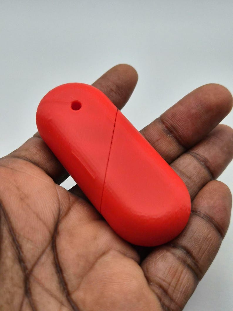 Puede incluir: Un objeto rojo brillante, de forma oblonga, con un peque&ntilde;o agujero en un extremo y una l&iacute;nea diagonal en la superficie. El objeto se sostiene en una mano de piel oscura, sobre un fondo blanco.