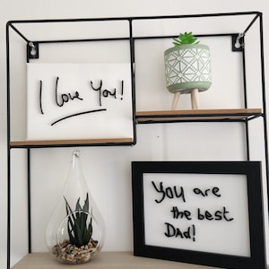 Op de afbeelding: Een zwarte metalen plank met drie planken. De bovenste plank heeft een kleine groene plant in een pot met een wit en groen geometrisch patroon. De middelste plank heeft een wit canvas met de woorden "Ik hou van je!" geschreven in zwarte stift. De onderste plank heeft een heldere glazen traanvormige vaas met een kleine groene plant en een kleine stapel stenen. De rechterkant van de onderste plank heeft een zwart ingelijst droogwisbord met de woorden "Je bent de beste papa!" geschreven in zwarte stift.