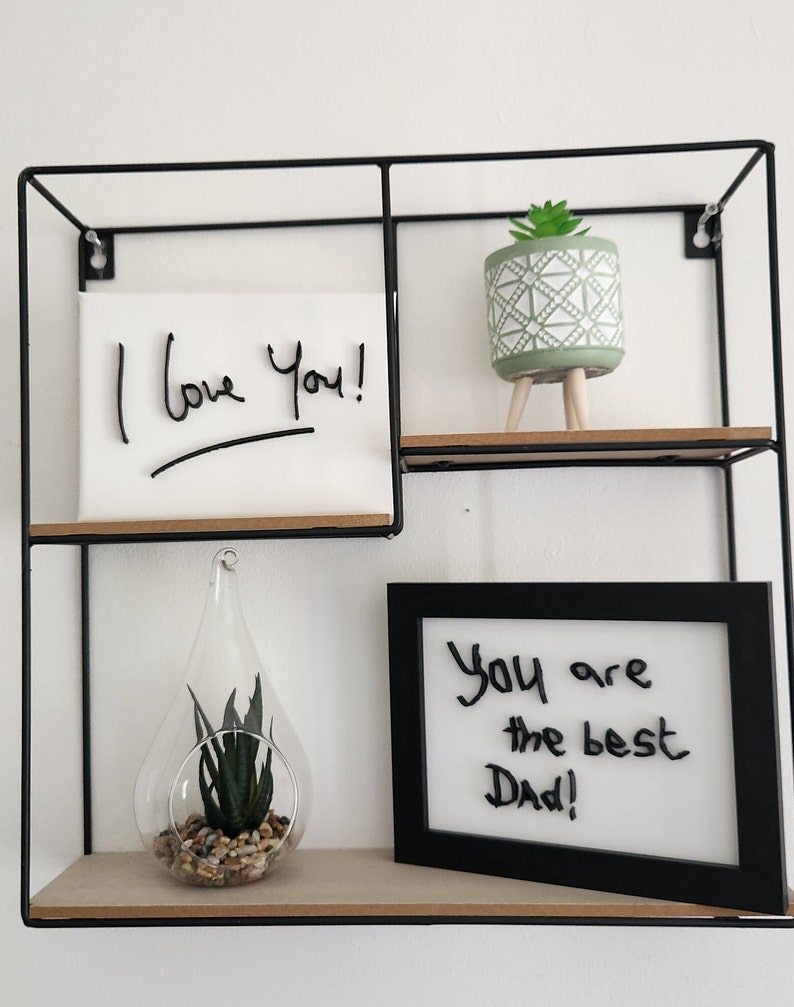 Op de afbeelding: Een zwarte metalen plank met drie planken. De bovenste plank heeft een kleine groene plant in een pot met een wit en groen geometrisch patroon. De middelste plank heeft een wit canvas met de woorden "Ik hou van je!" geschreven in zwarte stift. De onderste plank heeft een heldere glazen traanvormige vaas met een kleine groene plant en een kleine stapel stenen. De rechterkant van de onderste plank heeft een zwart ingelijst droogwisbord met de woorden "Je bent de beste papa!" geschreven in zwarte stift.