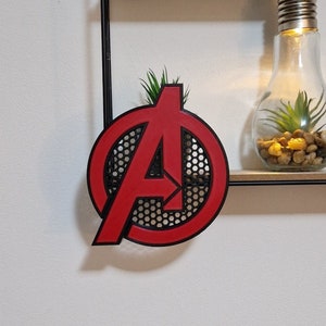 Op de afbeelding: Een rood en zwart Avengers-logo met een honingraatpatroon. Het logo is gemonteerd op een zwart rek met een kleine plant in een glazen pot.