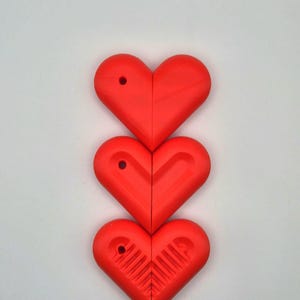 Puede incluir: Tres objetos rojos en forma de coraz&oacute;n dispuestos verticalmente sobre un fondo blanco. Cada coraz&oacute;n est&aacute; dividido por la mitad, con un peque&ntilde;o agujero cerca de la parte superior. El coraz&oacute;n inferior tiene un dise&ntilde;o texturizado en el interior.