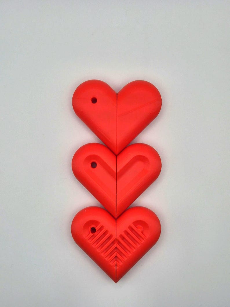 Puede incluir: Tres objetos rojos en forma de coraz&oacute;n dispuestos verticalmente sobre un fondo blanco. Cada coraz&oacute;n est&aacute; dividido por la mitad, con un peque&ntilde;o agujero cerca de la parte superior. El coraz&oacute;n inferior tiene un dise&ntilde;o texturizado en el interior.