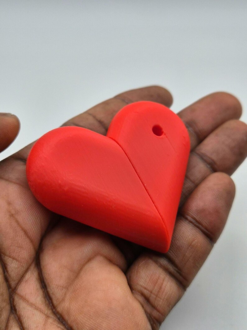 Puede incluir: Un objeto rojo brillante en forma de coraz&oacute;n con un agujero en la parte superior. El coraz&oacute;n est&aacute; sostenido en la mano de una persona. El objeto parece estar hecho de pl&aacute;stico o un material similar.