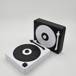 Può includere: Due giradischi in miniatura, uno bianco e uno nero, sono esposti su sfondo bianco. Il giradischi bianco ha un disco nero, e il giradischi nero ha un disco bianco. Entrambi hanno un braccio e una manopola di controllo.