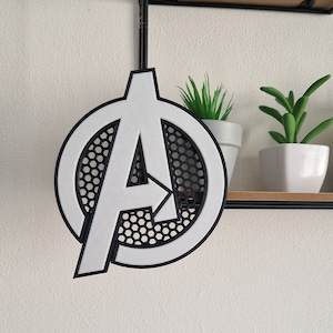 Op de afbeelding: Wit plastic Avengers-logo met een zwart honingraatpatroon in de cirkel. Het logo hangt aan een zwart touwtje.