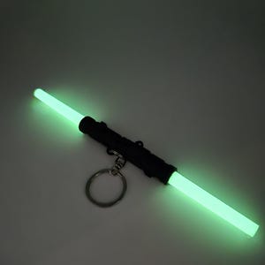 K&ouml;nnte beinhalten: Ein schwarzer doppelseitiger Lichtschwert-Schl&uuml;sselanh&auml;nger mit leuchtenden gr&uuml;nen Lichtr&ouml;hren. Der Schl&uuml;sselanh&auml;nger hat einen silbernen Ring zum Befestigen an Schl&uuml;sseln oder einer Tasche. Das Lichtschwert ist ein Spielzeug oder ein Neuheitenartikel.