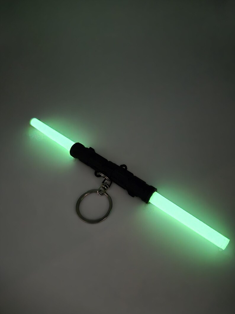 K&ouml;nnte beinhalten: Ein schwarzer doppelseitiger Lichtschwert-Schl&uuml;sselanh&auml;nger mit leuchtenden gr&uuml;nen Lichtr&ouml;hren. Der Schl&uuml;sselanh&auml;nger hat einen silbernen Ring zum Befestigen an Schl&uuml;sseln oder einer Tasche. Das Lichtschwert ist ein Spielzeug oder ein Neuheitenartikel.