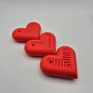 Puede incluir: Tres objetos rojos en forma de coraz&oacute;n con un dise&ntilde;o suave y redondeado. Cada coraz&oacute;n tiene un peque&ntilde;o agujero en la parte superior y uno tiene un patr&oacute;n texturizado en la mitad inferior. Los objetos est&aacute;n dispuestos sobre un fondo blanco.