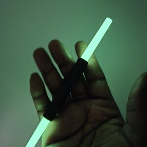 K&ouml;nnte beinhalten: Ein leuchtender, neongr&uuml;ner Stab mit einem schwarzen Griff. Der Stab wird in einer Hand vor einem gr&uuml;nen Hintergrund gehalten. Der neongr&uuml;ne Stab ist rechteckig und strahlt helles Licht aus.