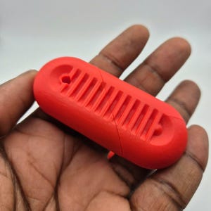 Puede incluir: Un objeto oblongo de color rojo brillante con una serie de ranuras paralelas y un extremo curvo. El objeto, sostenido en una mano, parece estar hecho de un material s&oacute;lido y tiene una grieta. Las ranuras son uniformes.