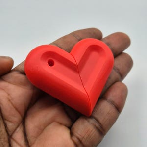 Puede incluir: Un objeto rojo brillante en forma de coraz&oacute;n con un agujero, sostenido en una mano. El coraz&oacute;n est&aacute; dividido por la mitad. El objeto parece estar hecho de un material s&oacute;lido, posiblemente pl&aacute;stico o resina. El fondo es blanco liso.