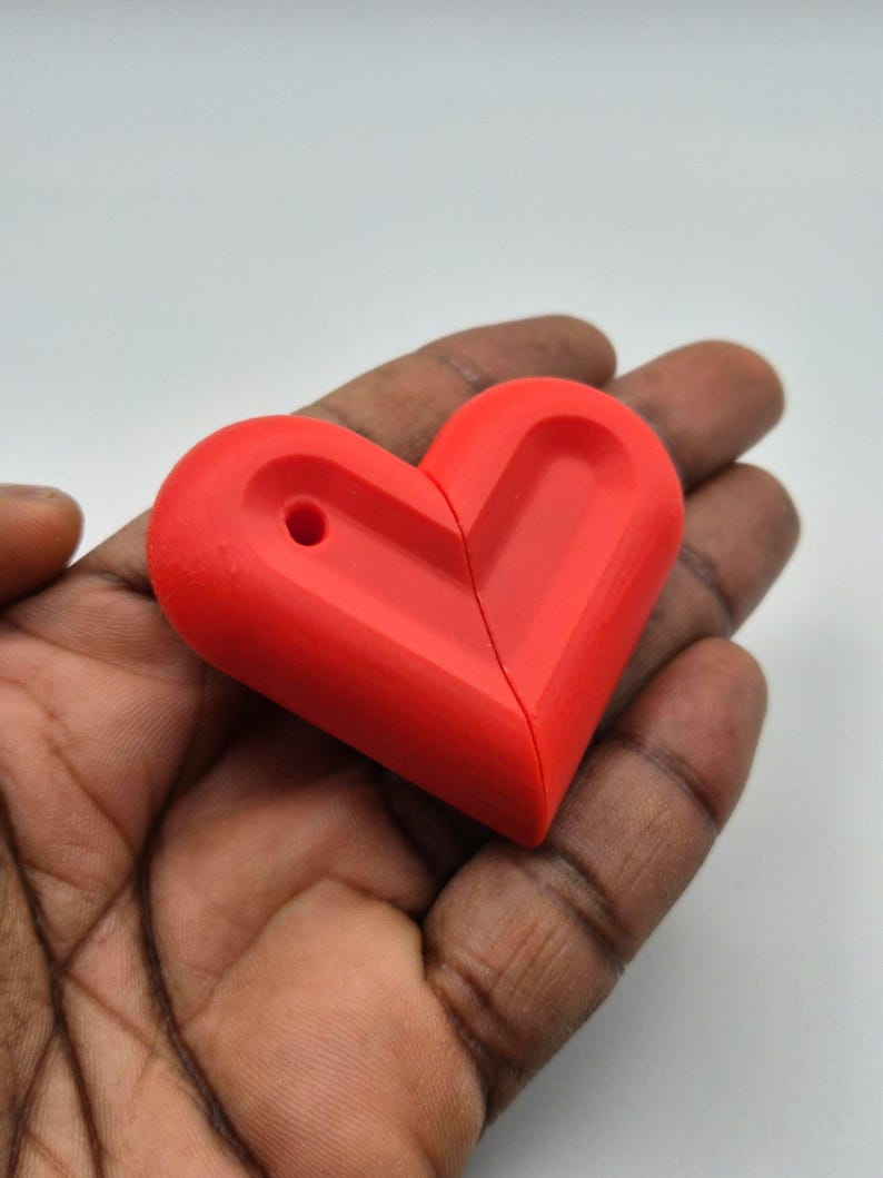 Puede incluir: Un objeto rojo brillante en forma de coraz&oacute;n con un agujero, sostenido en una mano. El coraz&oacute;n est&aacute; dividido por la mitad. El objeto parece estar hecho de un material s&oacute;lido, posiblemente pl&aacute;stico o resina. El fondo es blanco liso.