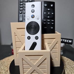 Puede incluir: Un soporte de madera beige para mandos a distancia con tres ranuras. El soporte tiene forma de caja y tiene una base negra. Hay tres mandos a distancia en el soporte, uno blanco y dos negros.