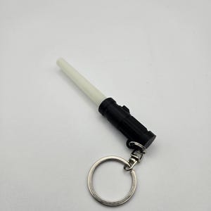 K&ouml;nnte beinhalten: Ein schwarz-wei&szlig;es Lichtschwert-Schl&uuml;sselanh&auml;nger. Der Schl&uuml;sselanh&auml;nger hat einen schwarzen Griff, eine wei&szlig;e Klinge und einen silbernen Schl&uuml;sselring. Das Lichtschwert ist eine Miniatur-Replik der ikonischen Waffe aus der Science-Fiction-Reihe.