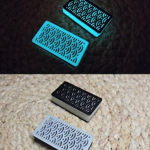 May include: Two small rectangular boxes with a black and white geometric pattern. The boxes are made of a material that glows in the dark. The boxes are on a woven surface.