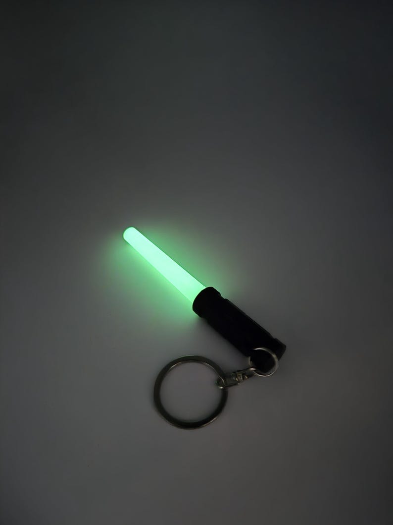 K&ouml;nnte beinhalten: Ein leuchtend gr&uuml;nes Schl&uuml;sselanh&auml;ngerlicht mit einem schwarzen Griff und einem silbernen Schl&uuml;sselring. Das Licht strahlt gleichm&auml;&szlig;ig hell, und der Griff ist zylindrisch. Der Schl&uuml;sselring ist mit einer kleinen Metallschlaufe am Griff befestigt.