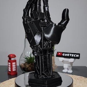 Op de afbeelding: Een zwarte 3D-geprinte robotarm met ingewikkelde details, staande op een zwarte ronde basis met een reflecterend oppervlak. De hand is omhoog geheven met gestrekte vingers, wat lijkt op een begroetings- of aanbiedingsgebaar.