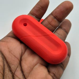 Puede incluir: Memoria USB roja brillante con forma rectangular redondeada. La unidad tiene un peque&ntilde;o agujero en un extremo y una superficie texturizada. El dispositivo se sostiene en una mano sobre un fondo blanco.