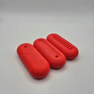 Puede incluir: Tres objetos oblongos de color rojo brillante. Los objetos tienen un dise&ntilde;o liso y redondeado con un peque&ntilde;o agujero en un extremo. Un objeto tiene una serie de l&iacute;neas paralelas en su superficie superior. Los objetos est&aacute;n dispuestos sobre un fondo blanco.