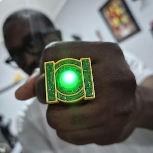 May include: A square-shaped ring featuring a bright green glowing circular center set within a textured green frame and a yellow border. The ring displays the iconic Green Lantern symbol design with a vibrant light emitting from the middle.