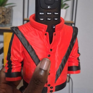 Peut inclure: Un support de télécommande décoratif rouge et noir en forme de veste rouge avec des rayures noires. La télécommande est noire et comporte plusieurs boutons de contrôle multimédia.