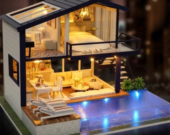 May include: A miniature dollhouse with a white exterior and a black roof. The dollhouse has a swimming pool, a balcony, and a kitchen. The dollhouse is lit up with lights.
