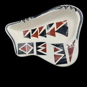 May include: A white ceramic ashtray with a black and red geometric pattern. The ashtray has a unique shape with a scalloped edge and a black border. The design features triangles and diamond shapes.