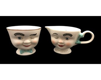 Puede incluir: Un juego de dos tazas de cerámica blanca con caras guiñando un ojo. Las tazas presentan el logotipo de Bailey's y un lazo azul.