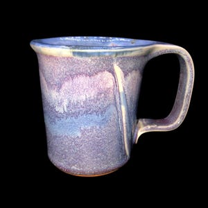 May include: A handcrafted ceramic mug with an unusual, organic form. The mug displays a mix of blue, purple, and white tones, with a textured finish. The handle is a curved, robust design. The mug's base is a warm brown colour.