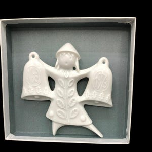 Puede incluir: Adorno navideño de porcelana blanca con una niña que sostiene dos campanas. Las campanas están decoradas con los años 1972 y 1973.