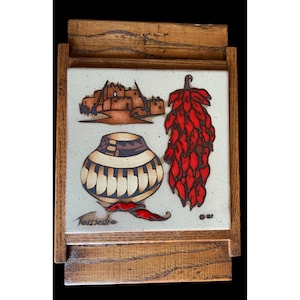 May include: A wooden framed tile with a Southwestern design. The tile features a red chili pepper, a brown pot with a geometric pattern, and a depiction of adobe buildings. The tile is signed "Telssedre" in the bottom left corner.