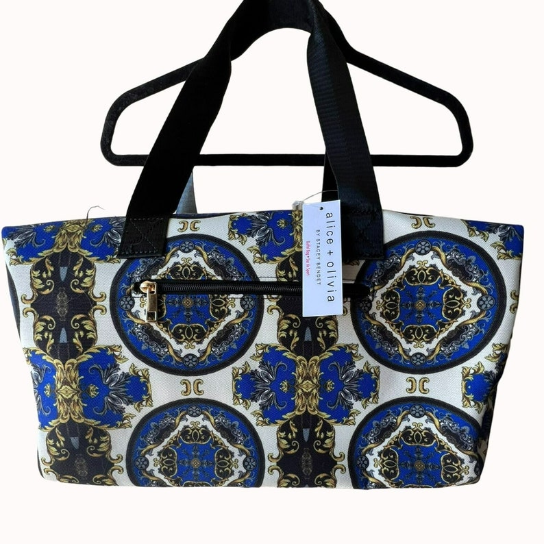 Puede incluir: Una bolsa de mano blanca con un dise&ntilde;o estampado en azul y dorado. La bolsa tiene asas negras y cierre de cremallera. La bolsa tiene una etiqueta que dice "alice + olivia by Stacey Bendet" y "Love, Alice + Olivia".