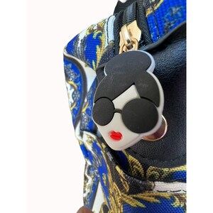 Puede incluir: Una bolsa negra con cremallera con un dise&ntilde;o estampado en azul y dorado. El tirador de la cremallera es una cara de dibujos animados blanca con gafas de sol negras y labios rojos.