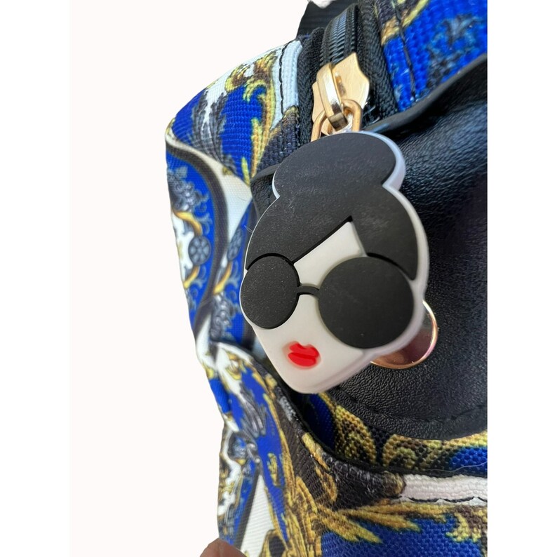 Puede incluir: Una bolsa negra con cremallera con un dise&ntilde;o estampado en azul y dorado. El tirador de la cremallera es una cara de dibujos animados blanca con gafas de sol negras y labios rojos.