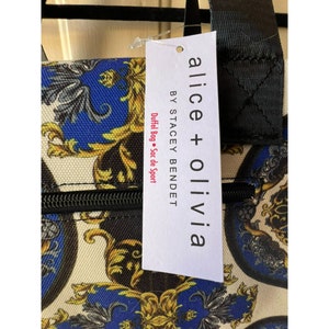 Puede incluir: Una bolsa de lona blanca y negra con cierre de cremallera. La bolsa tiene un dise&ntilde;o estampado en dorado y azul con una etiqueta que dice "alice + olivia BY STACEY BENDEt DUFFel Bag &bull; Sac de Sport".