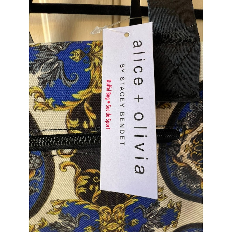 Puede incluir: Una bolsa de lona blanca y negra con cierre de cremallera. La bolsa tiene un dise&ntilde;o estampado en dorado y azul con una etiqueta que dice "alice + olivia BY STACEY BENDEt DUFFel Bag &bull; Sac de Sport".