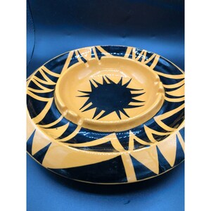 May include: A ceramic ashtray with a black and orange geometric design. The ashtray has a central well with a black starburst pattern.