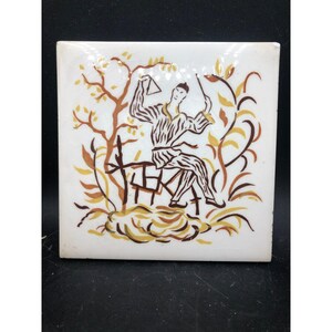 May include: Square ceramic tile with a hand-painted design. The tile features a seated figure in brown and white striped clothing, holding a triangle and a stick. The background includes brown and yellow stylized trees and foliage.