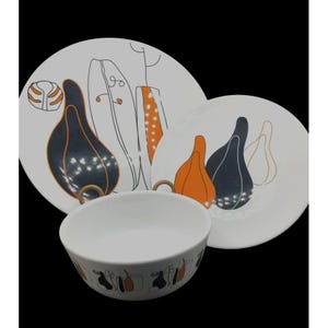 Peut inclure: Un service de vaisselle en céramique blanche comprenant un bol, une petite assiette et une grande assiette. Les assiettes présentent un motif de formes abstraites orange et bleu marine. Le bol a une anse dorée et un motif répété des mêmes formes.