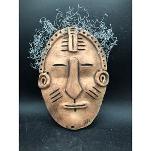 May include: A tan-colored ceramic mask with a stylized face, featuring closed eyes, a prominent nose, and a small mouth. The mask has wire hair and decorative markings. The mask is likely for wall decor.