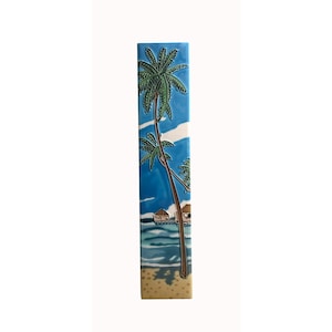 Op de afbeelding: Een rechthoekige keramische tegel met een tropische strandscène. Het kunstwerk toont een blauwe lucht met witte wolken, een palmboom en een strand met hutten. De tegel is ongeveer 76 cm hoog.