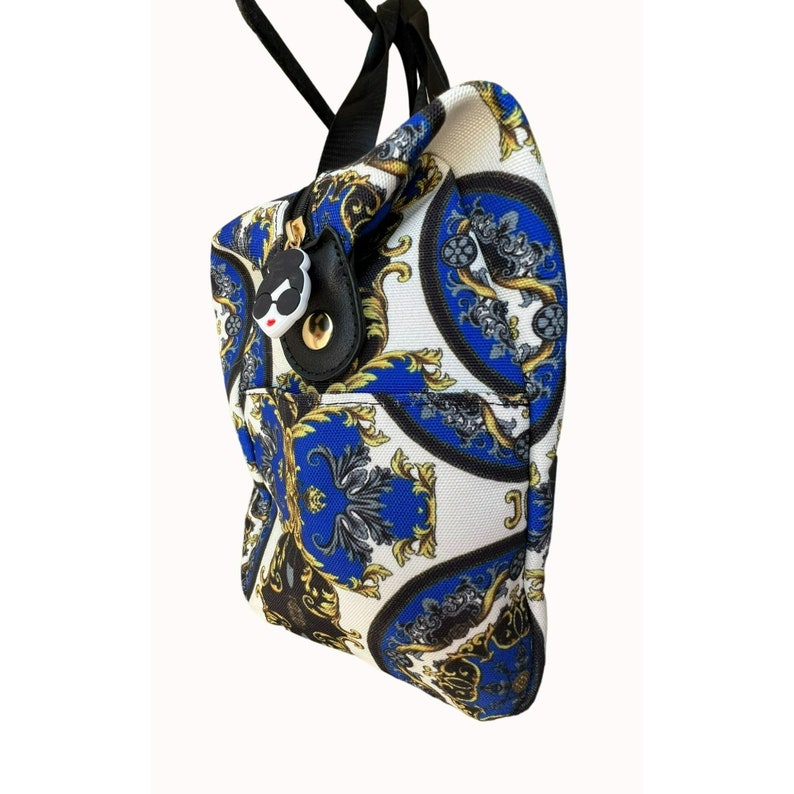 Puede incluir: Una bolsa de lona blanca con un patr&oacute;n barroco azul y dorado. La bolsa tiene una correa negra y un peque&ntilde;o amuleto blanco y negro unido a la cremallera.