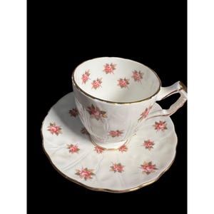 May include: A white teacup and saucer with a delicate floral pattern of pink roses and green leaves. The cup and saucer have a gold trim around the edges.