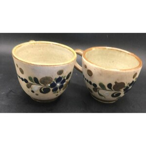May include: Two ceramic coffee mugs with brown, blue, and green floral designs. The mugs have a light brown glaze and a white background.