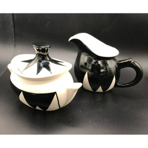 May include: A black and white ceramic creamer and sugar bowl set with a geometric design. The creamer has a white spout and handle. The sugar bowl has a lid with a black knob.