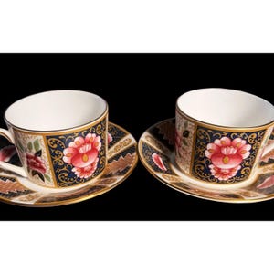 Könnte beinhalten: Zwei Teetassen und Untertassen mit einem Blumenmuster. Die Tassen und Untertassen sind weiß mit einem blauen und goldenen Rand. Das Blumenmuster zeigt rosa, rote und weiße Blumen.