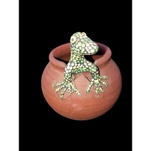 May include: A small terracotta pot with a green and black spotted lizard figurine emerging from the top. The lizard has yellow eyes and is perched on the rim of the pot.